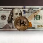 Close-up of a Bitcoin coin placed in front of a US hundred-dollar bill, highlighting cryptocurrency versus traditional money.