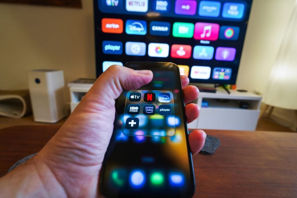 Close-up of a hand holding a phone displaying streaming apps in front of a TV with multiple app icons.