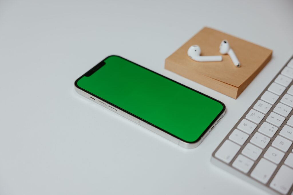 A white smartphone with a green screen next to wireless earphones and a keyboard on a white surface.