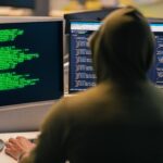 A person in a hoodie coding on dual monitors, depicting cybersecurity and hacking themes.