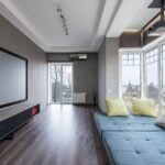 Comfortable settee with colorful cushions placed near big windows against contemporary television set in spacious room of modern stylish apartment